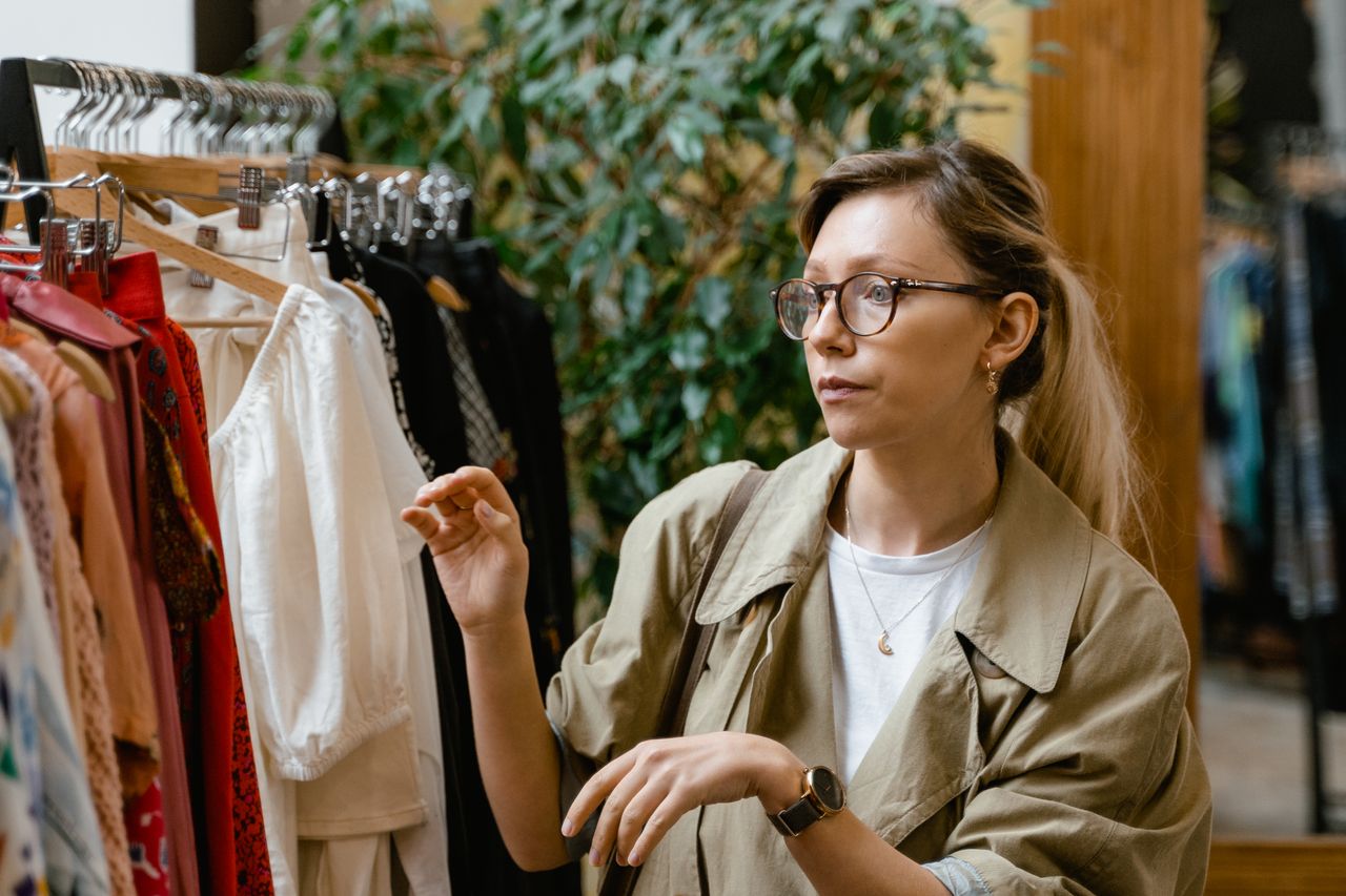 Stylish woman with glasses shopping for clothes at an indoor boutique, browsing clothing racks.