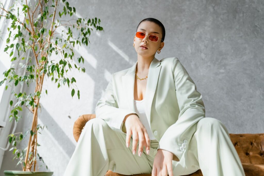 Confident woman in a stylish suit and sunglasses, posing indoors with natural sunlight.